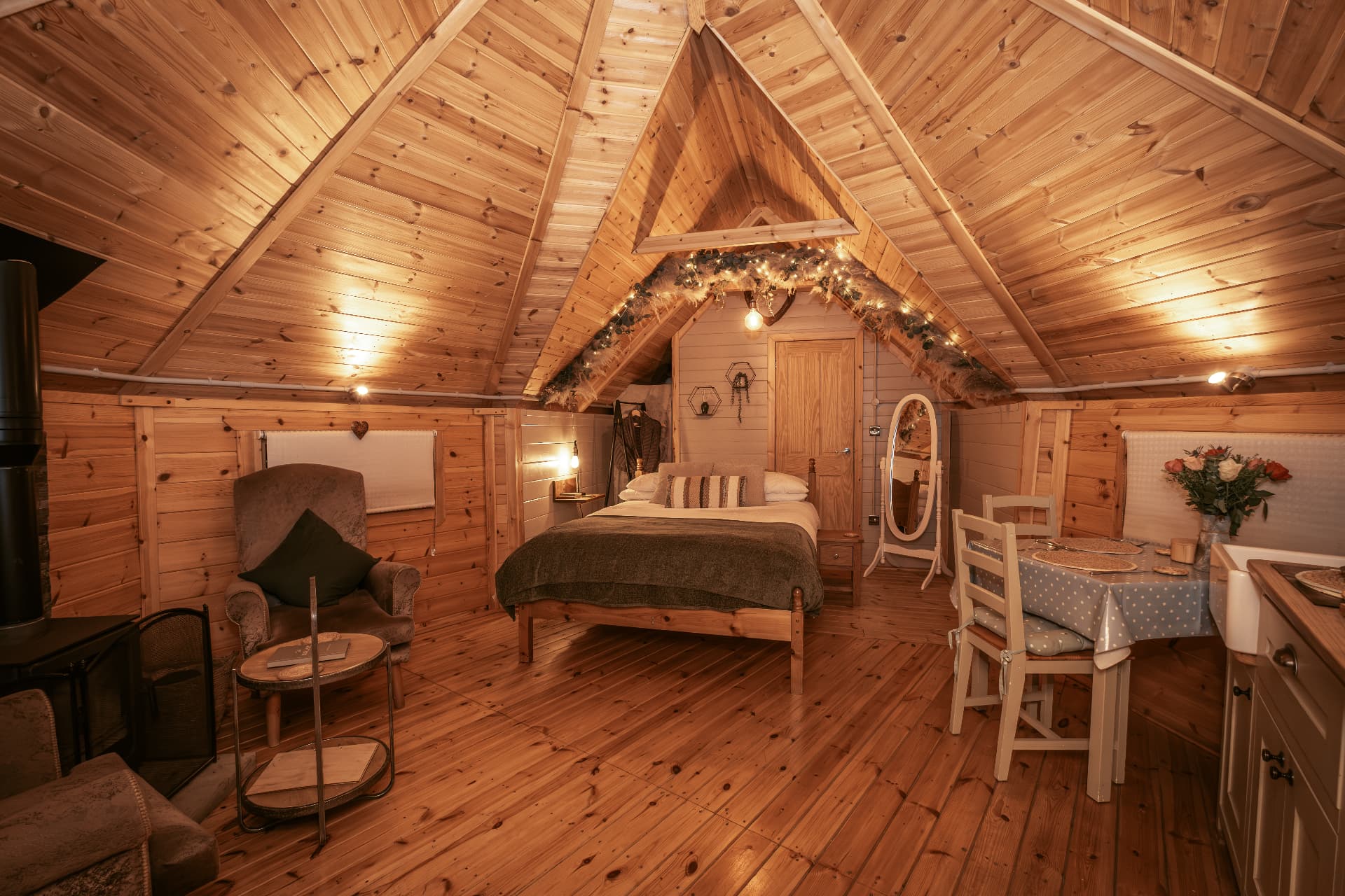 The full interior of The Mallard — wood burner, armchair, bed and dining table beneath the pitched ceiling